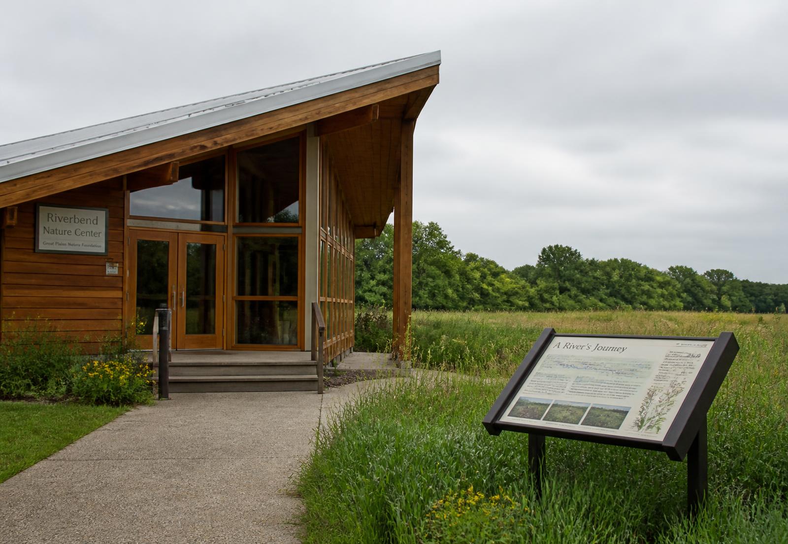 A timber-and-glass nature center building at the edge of a meadow with an interpretive sign nearby.